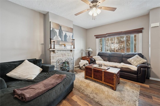 living room with baseboards, dark wood-type flooring, a textured ceiling, ceiling fan, and a stone fireplace