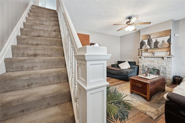 staircase featuring a textured ceiling, ceiling fan, a stone fireplace, and wood finished floors