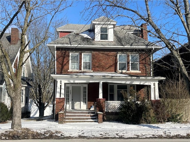 american foursquare style home with covered porch, a shingled roof, and brick siding