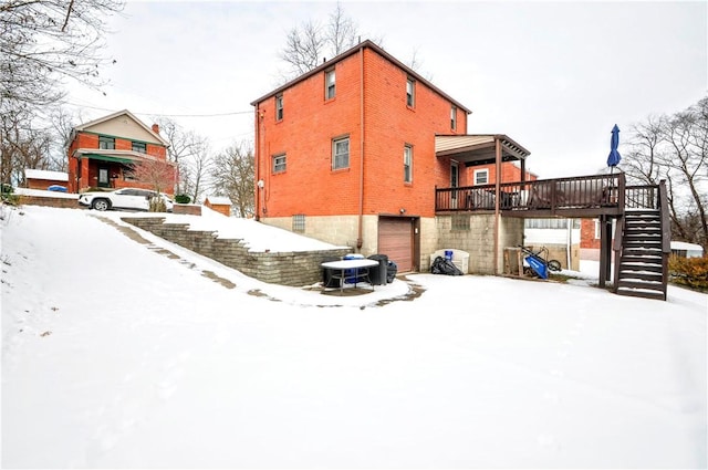 snow covered house featuring a garage, a wooden deck, stairs, and brick siding