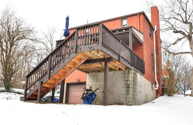 snow covered house with a garage, brick siding, a chimney, and stairs