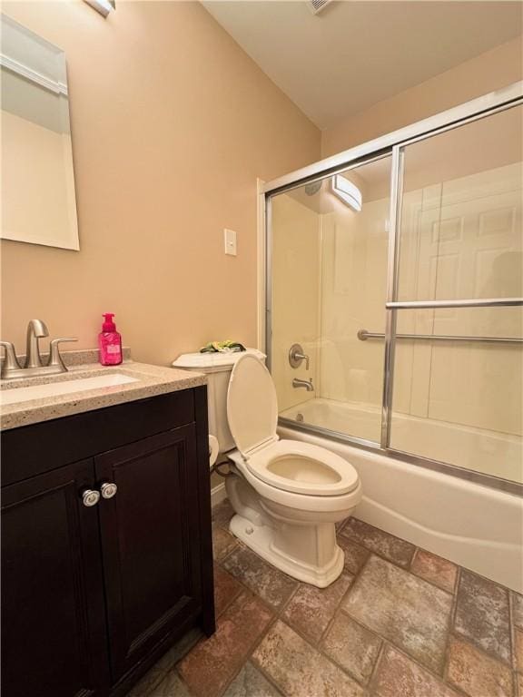 bathroom with stone finish floor, shower / bath combination with glass door, vanity, and toilet