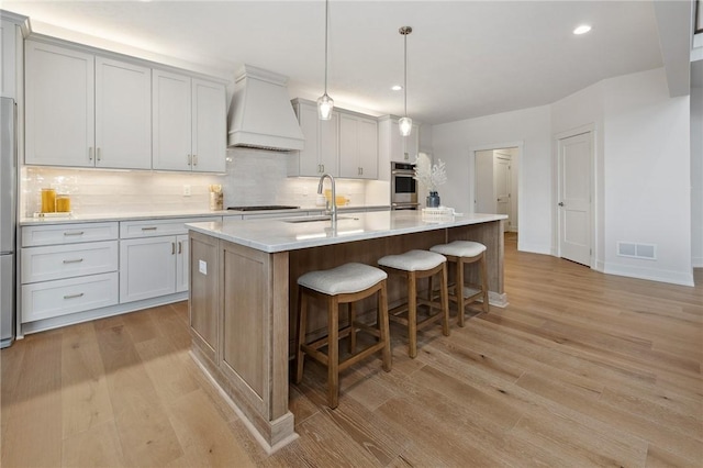 kitchen with a sink, light wood-style flooring, custom exhaust hood, visible vents, and decorative backsplash