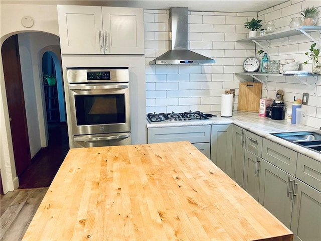 kitchen featuring open shelves, decorative backsplash, appliances with stainless steel finishes, butcher block countertops, and wall chimney exhaust hood