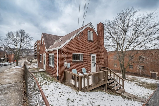 snow covered back of property featuring brick siding, roof with shingles, a chimney, fence, and a deck