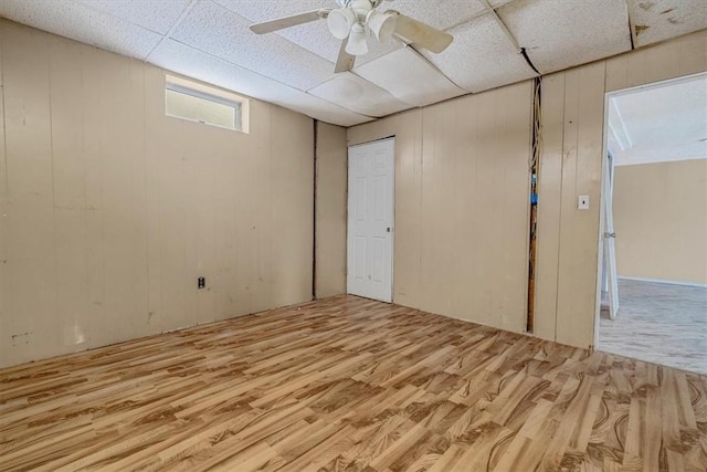 basement featuring ceiling fan, wooden walls, a drop ceiling, and light wood-style floors