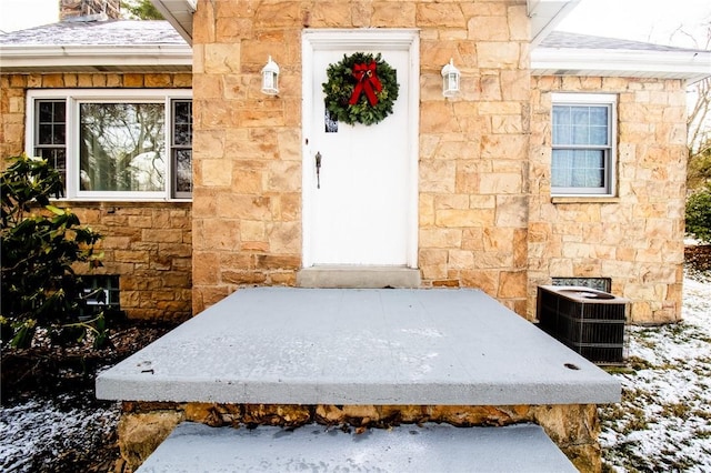 snow covered property entrance featuring stone siding, central AC, and roof with shingles