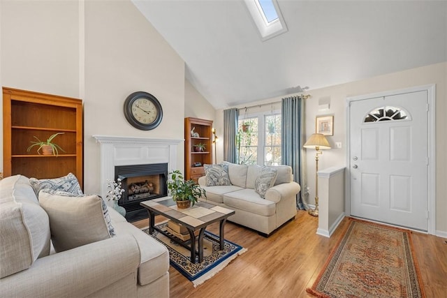 living room featuring a skylight, light wood-style floors, a fireplace with flush hearth, high vaulted ceiling, and baseboards