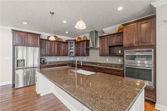 kitchen with glass insert cabinets, stainless steel appliances, dark brown cabinets, wall chimney range hood, and a sink