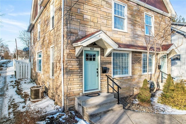 snow covered property entrance featuring stone siding, central AC, and fence