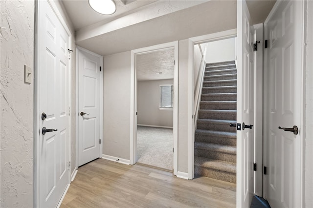 corridor with light wood-type flooring, stairway, baseboards, and a textured wall