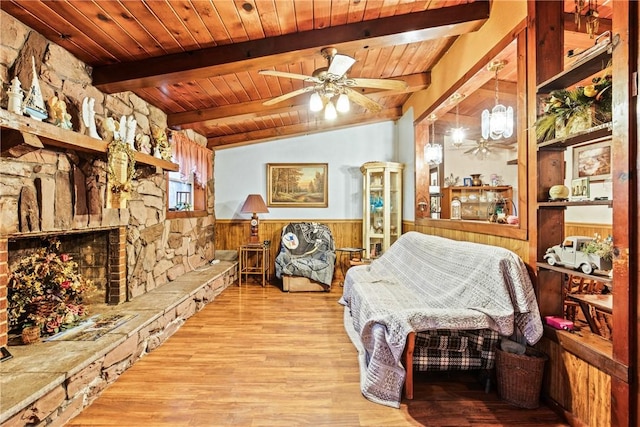 bedroom with light wood finished floors, wainscoting, wooden ceiling, vaulted ceiling with beams, and a stone fireplace