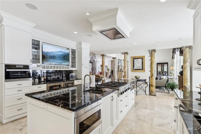 kitchen featuring decorative columns, glass insert cabinets, appliances with stainless steel finishes, white cabinetry, and an island with sink