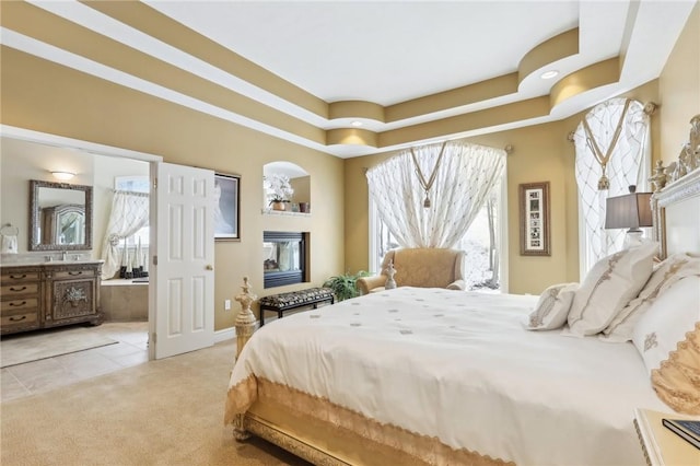 bedroom featuring a raised ceiling, connected bathroom, light colored carpet, a fireplace, and light tile patterned flooring