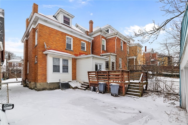 snow covered property with a deck, brick siding, a chimney, and central AC unit