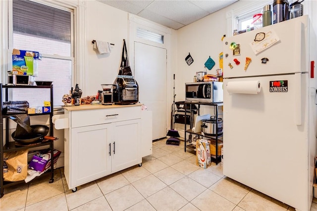 kitchen featuring stainless steel microwave, freestanding refrigerator, light tile patterned flooring, white cabinetry, and a drop ceiling