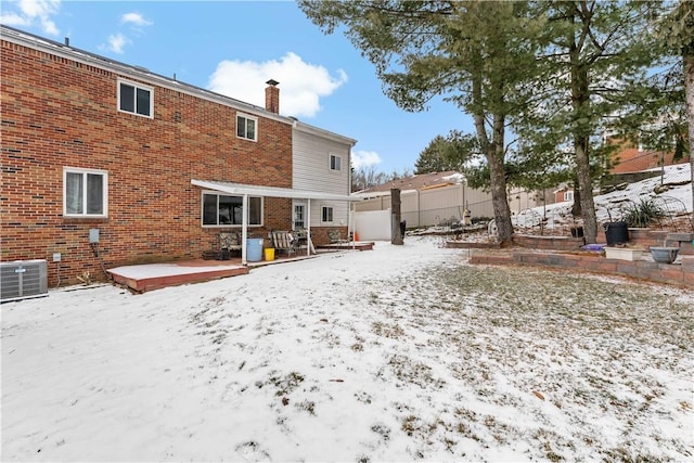 snow covered rear of property with brick siding, a chimney, central AC, fence, and a deck