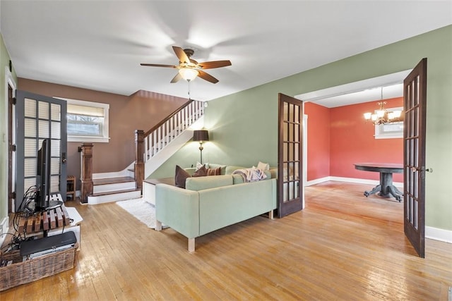 living area featuring stairs, french doors, light wood-style flooring, baseboards, and ceiling fan with notable chandelier