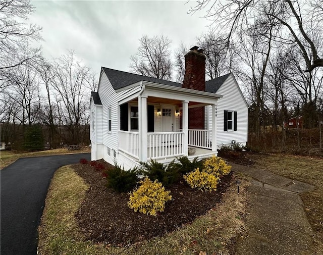 bungalow-style house with aphalt driveway, covered porch, a shingled roof, and a chimney