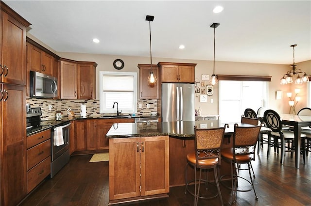 kitchen featuring hanging light fixtures, appliances with stainless steel finishes, brown cabinetry, a sink, and a kitchen island