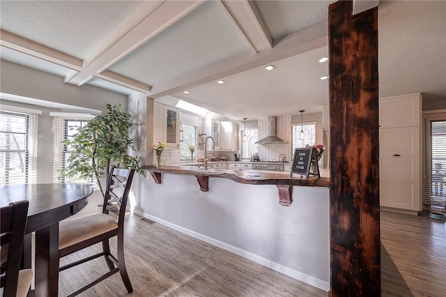 kitchen featuring a peninsula, white cabinetry, wall chimney range hood, beam ceiling, and decorative light fixtures