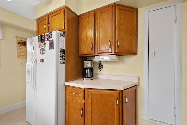 kitchen featuring white refrigerator with ice dispenser, light countertops, brown cabinetry, light tile patterned flooring, and baseboards