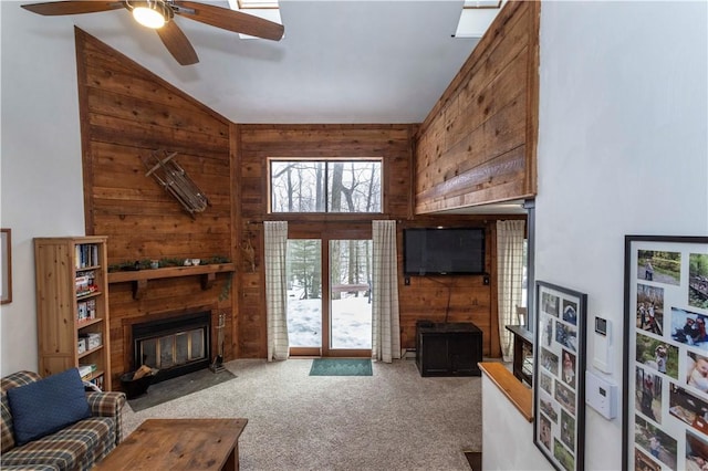 living room with high vaulted ceiling, wooden walls, a fireplace with flush hearth, carpet flooring, and a ceiling fan