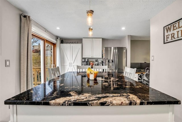 kitchen with a textured ceiling, white cabinetry, stainless steel fridge with ice dispenser, dark stone counters, and decorative light fixtures