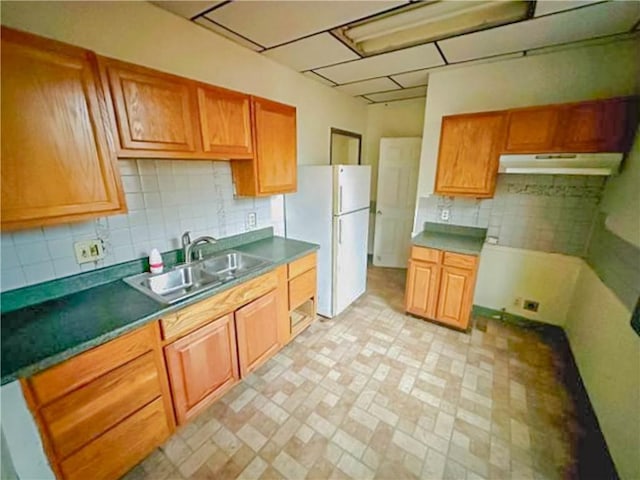 kitchen featuring under cabinet range hood, a sink, backsplash, freestanding refrigerator, and brown cabinetry