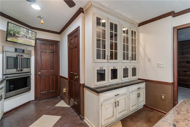 kitchen featuring crown molding, double oven, glass insert cabinets, wainscoting, and dark stone countertops