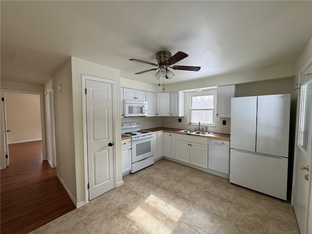 kitchen with white appliances, a sink, a ceiling fan, baseboards, and white cabinets