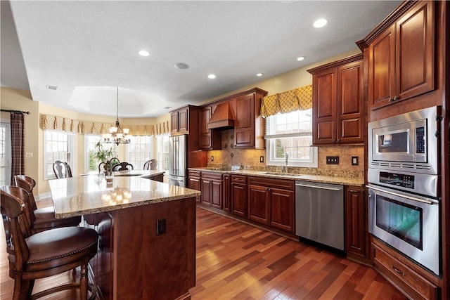 kitchen with a raised ceiling, custom range hood, an inviting chandelier, appliances with stainless steel finishes, and a sink