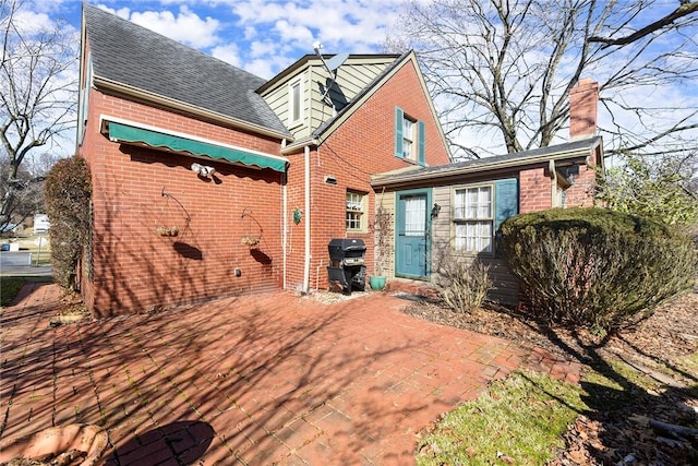 rear view of property featuring roof with shingles, brick siding, a chimney, and a patio area