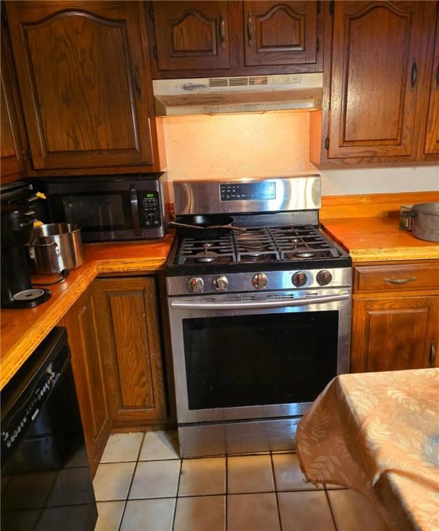 kitchen featuring light tile patterned floors, black dishwasher, stainless steel gas range oven, brown cabinets, and under cabinet range hood