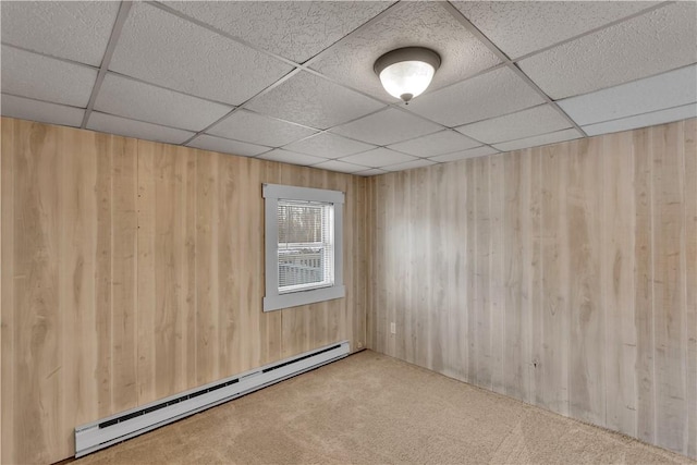 carpeted empty room with a drop ceiling, wood walls, and baseboard heating