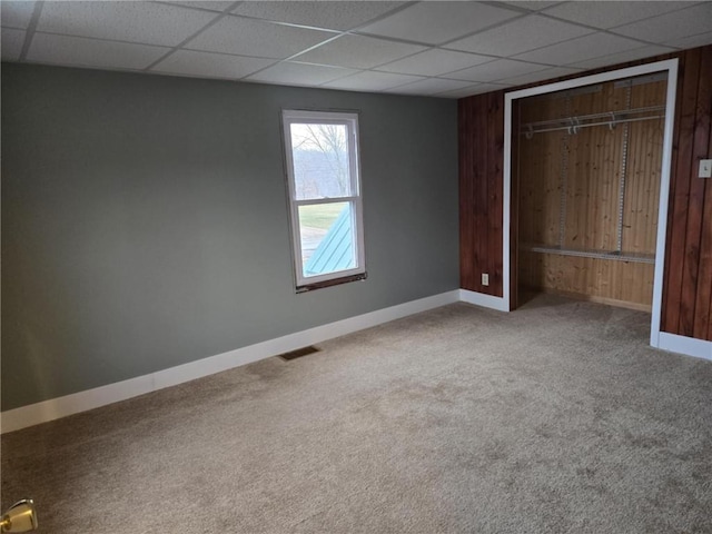 unfurnished bedroom featuring a closet, carpet flooring, wooden walls, a drop ceiling, and baseboards