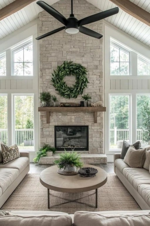 living room with a ceiling fan, beamed ceiling, wood finished floors, a stone fireplace, and high vaulted ceiling