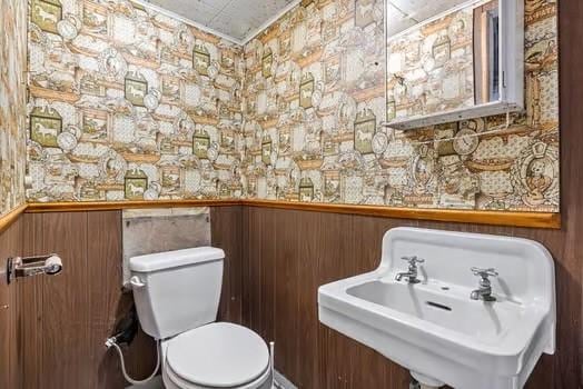 half bathroom with a wainscoted wall, a sink, toilet, and wallpapered walls