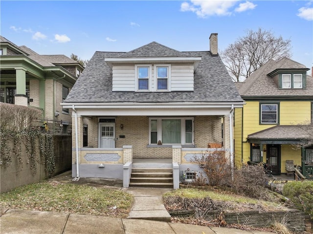 view of front of house with covered porch, brick siding, a shingled roof, fence, and a chimney