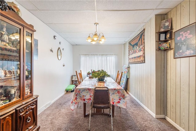 carpeted dining room with a chandelier, a drop ceiling, wood walls, and baseboards