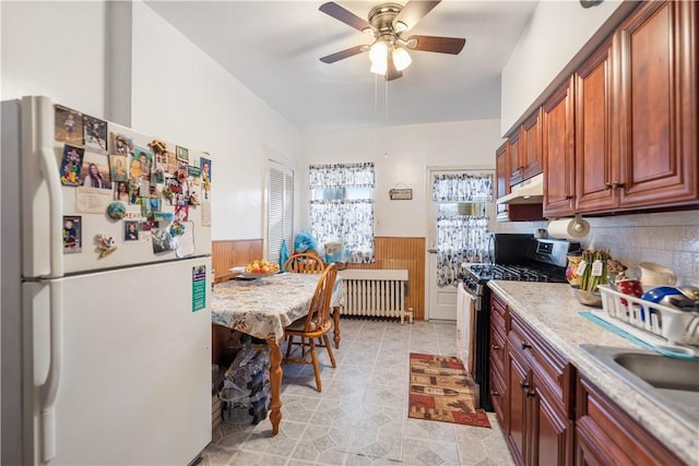 kitchen featuring a wainscoted wall, radiator, freestanding refrigerator, stainless steel gas stove, and under cabinet range hood