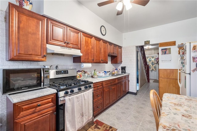 kitchen featuring stainless steel gas range oven, under cabinet range hood, a ceiling fan, freestanding refrigerator, and brown cabinets