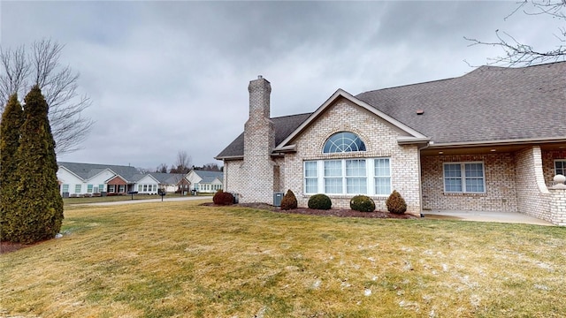 rear view of house featuring roof with shingles, brick siding, a chimney, a patio, and a lawn