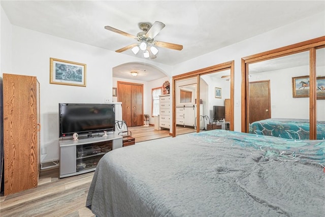 bedroom with light wood finished floors, ceiling fan, arched walkways, and two closets