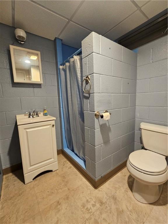 full bathroom with toilet, a sink, a shower with shower curtain, concrete block wall, and a paneled ceiling