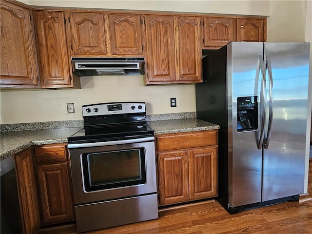 kitchen featuring brown cabinets, under cabinet range hood, dark countertops, dark wood finished floors, and appliances with stainless steel finishes