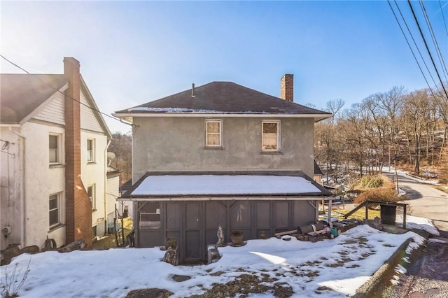 snow covered back of property featuring a garage, a chimney, a sunroom, and stucco siding