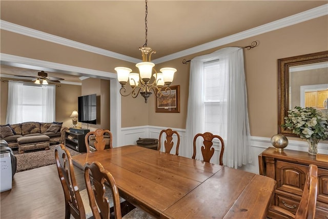 dining space featuring ornamental molding, ceiling fan with notable chandelier, a wainscoted wall, and wood finished floors