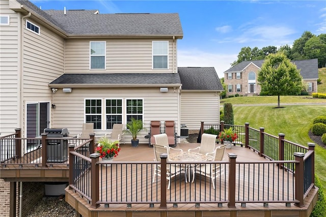 rear view of house with a shingled roof, outdoor dining area, a wooden deck, and a lawn