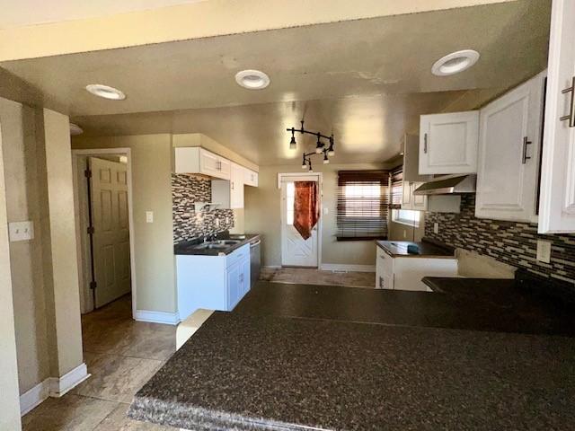 kitchen featuring baseboards, white cabinets, dark countertops, under cabinet range hood, and backsplash